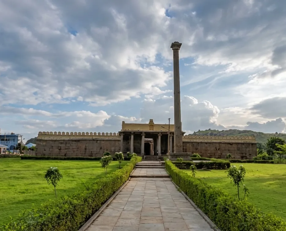 A temple at Thondanur (Thonnur) behind its long crenellated stone wall, a tall stone pillar standing before the pillared entrance porch, reached by a paved path between clipped hedges and lawns.
