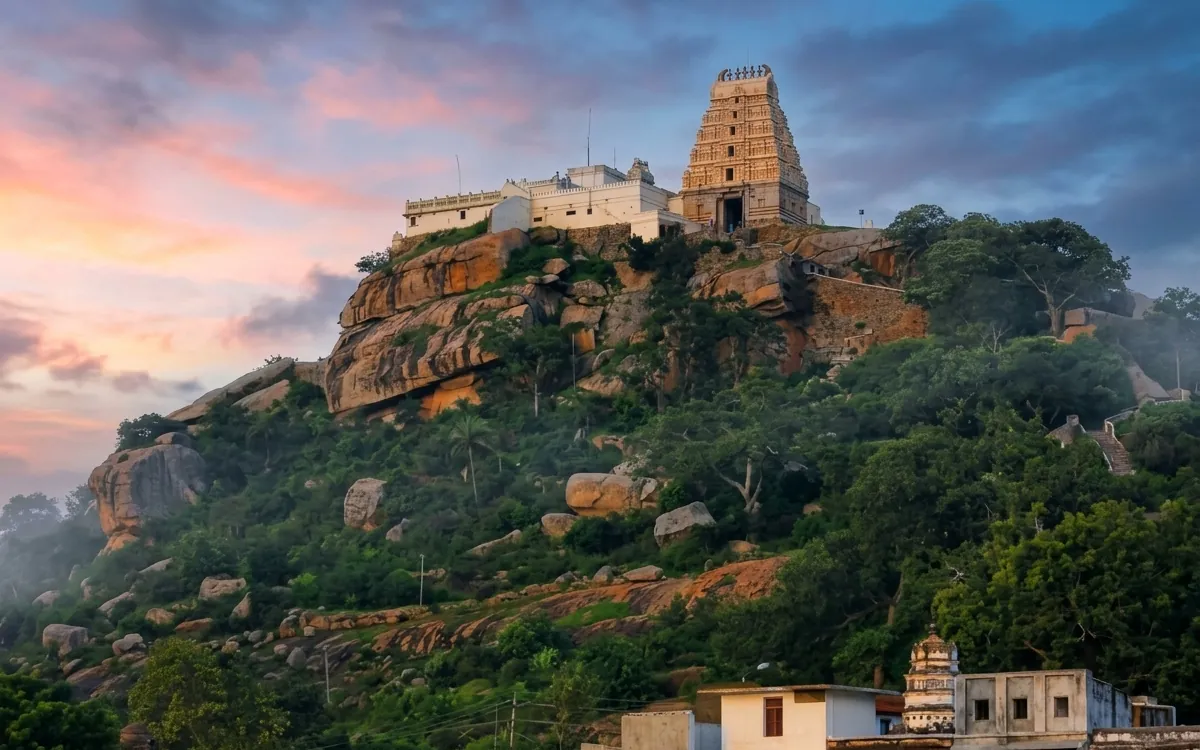 The Yoga Narasimha temple and its tall gopura on the summit of the rock above Melukote (Melkote) under a pink evening sky, the hillside below thick with trees and boulders, and an old pillared building of the town in the foreground.